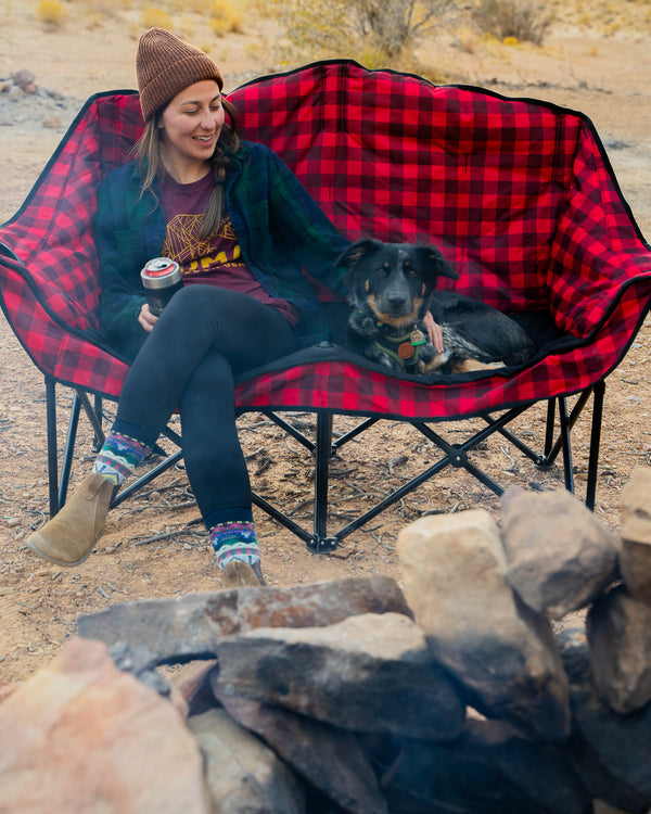 A woman sits in a red and black buffalo plaid camping chair with her dog resting beside her, holding a coffee cup in an outdoor desert setting near a fire pit.