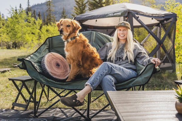 A woman sits in a green camping chair next to a golden retriever dog, with a canopy tent and outdoor forest background.