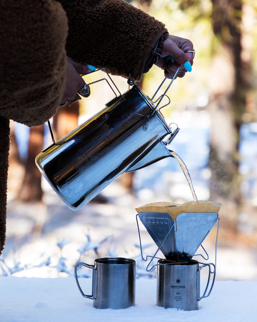 A person uses the Camping Coffee Set by Snow Peak to pour hot water from a metal kettle into a coffee dripper with a filter over a mug, making camp coffee outdoors among snowy trees.