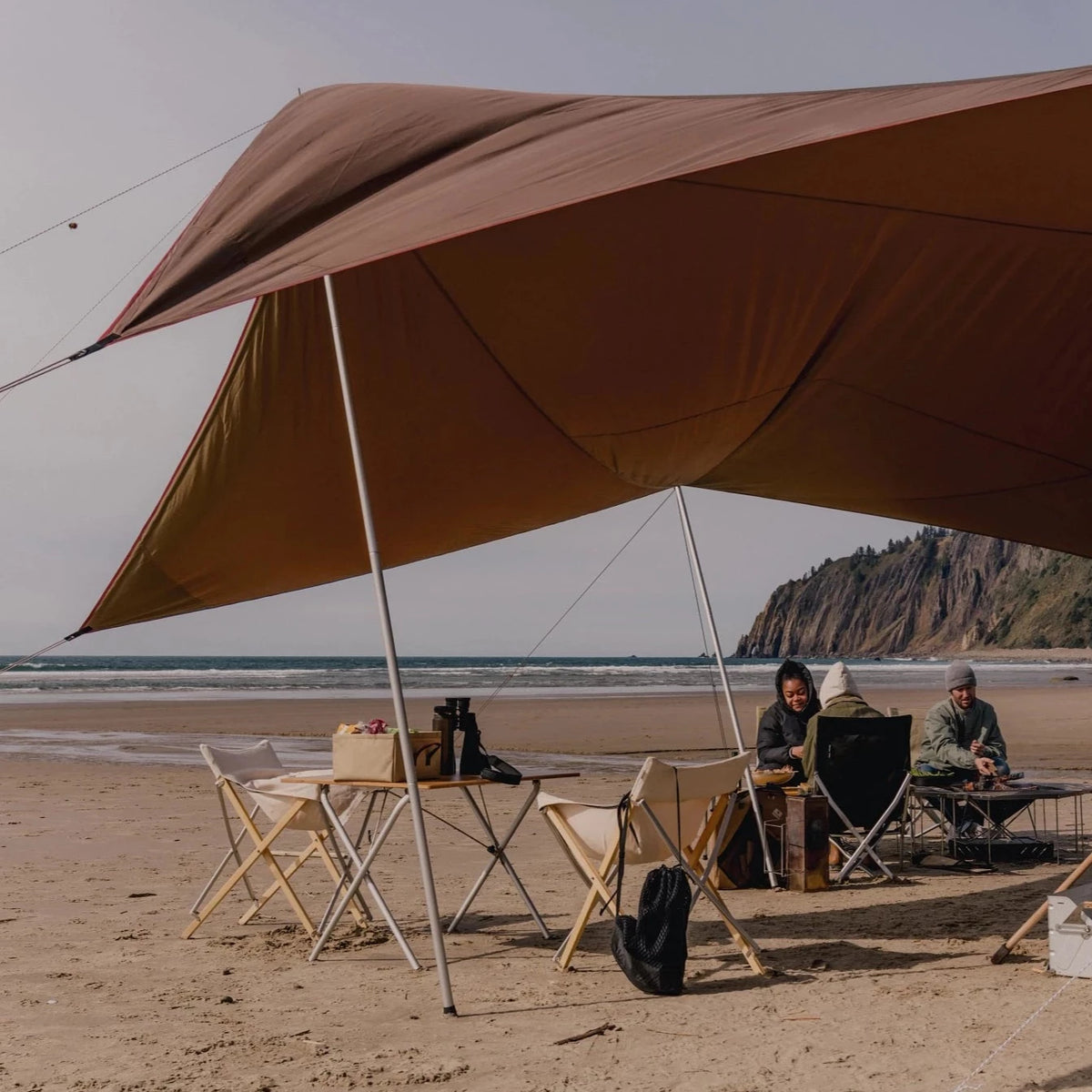 Two people sit under a large brown tarp canopy on a sandy beach, relaxing in Snow Peak Renewed Long Bamboo Chairs, surrounded by other camping gear, with cliffs and the sea in the background.
