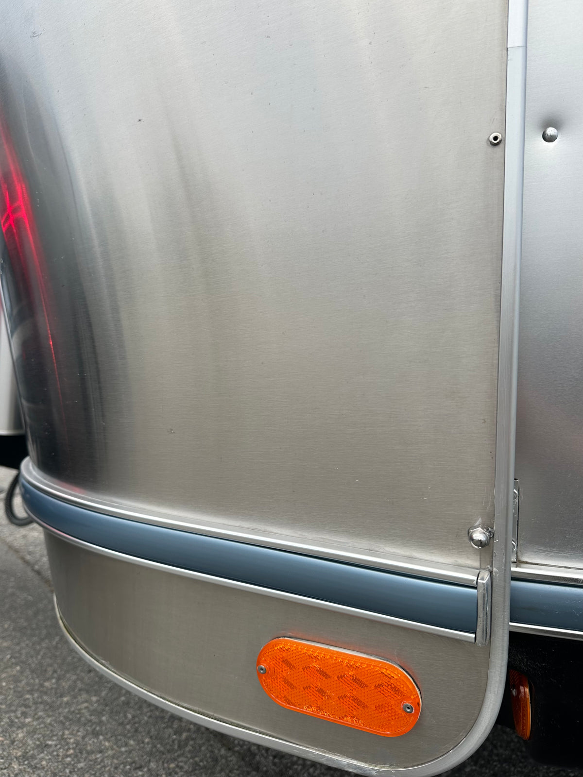 Close-up of a polished silver trailer featuring a visible seam, blue trim, and an orange reflector near the lower right corner—classic characteristics of riveted aluminum Airstream design. The blue trim highlights the Airstream Premium Replacement Rub Rail for Travel Trailers. Asphalt surface is visible in the background.