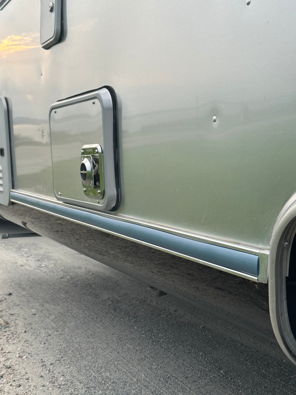 Close-up of the lower side of a light gray riveted aluminum Airstream, showing a small access panel and circular fitting, with part of a wheel well, pavement, and the Airstream Premium Replacement Rub Rail for Travel Trailers visible.