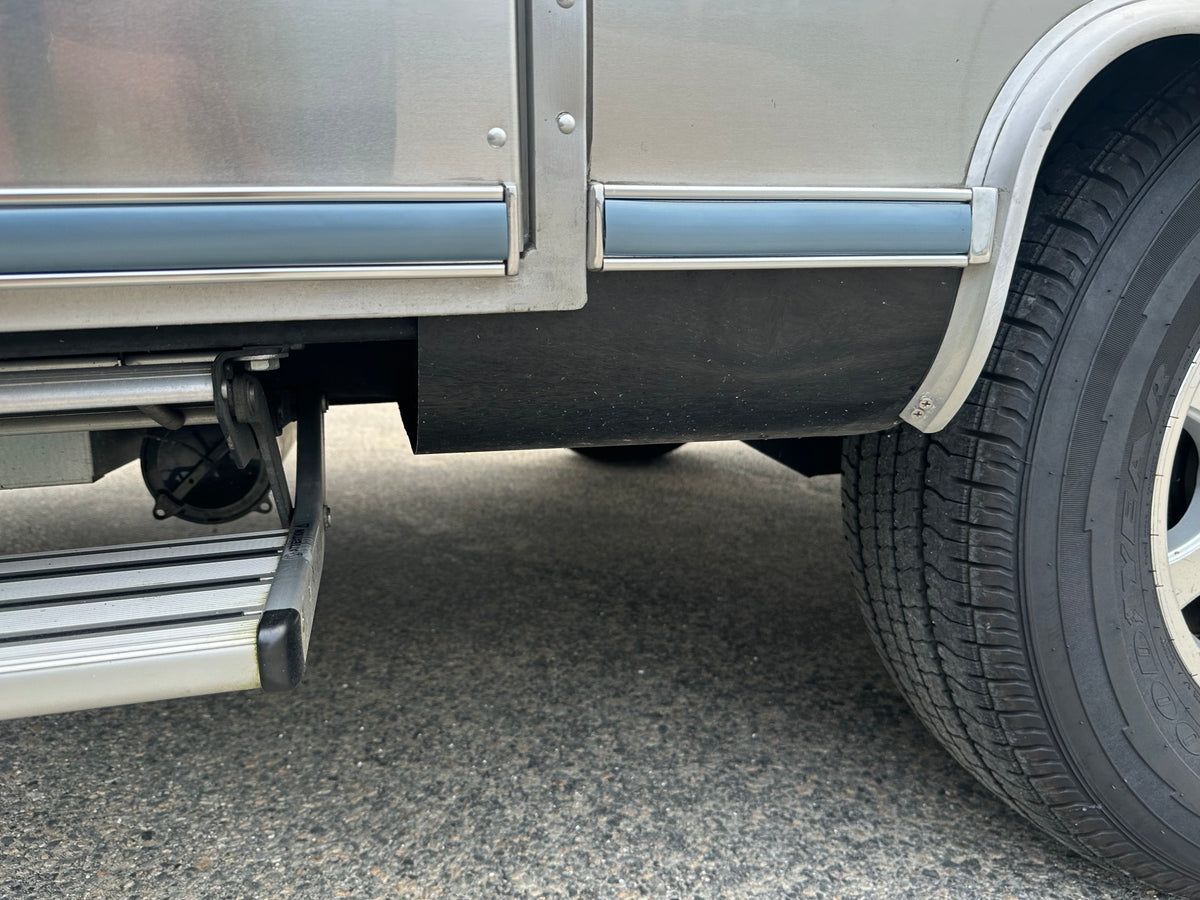 Close-up of a vehicle’s side showing a tire, metal running board step, and part of the lower body panel, featuring an Airstream Premium Replacement Rub Rail for Travel Trailers along the edge. The surface beneath is gray asphalt.