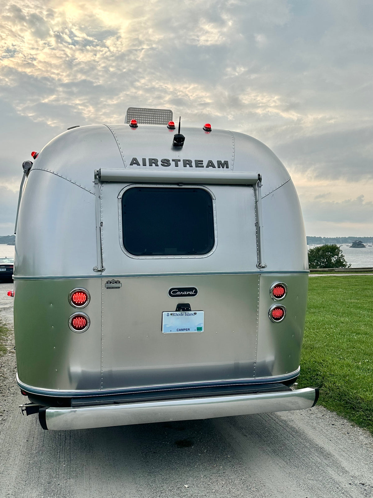 A silver Airstream trailer is parked near a grassy lakeside under a cloudy sky. The image captures the rear view of the riveted aluminum Airstream, showcasing its lights, license plate, and highlighting the Airstream Premium Replacement Rub Rail for Travel Trailers along the edge.