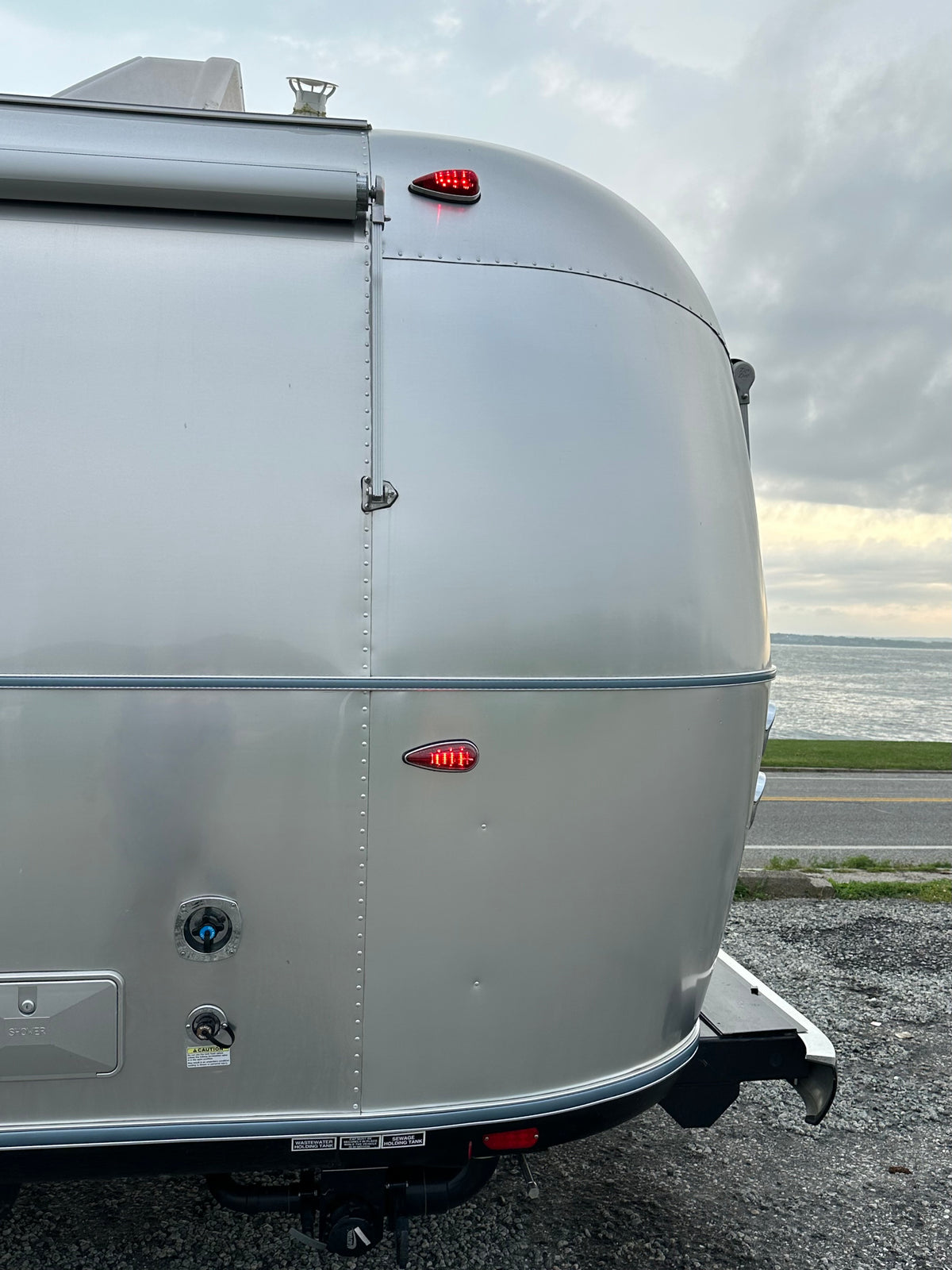 The rear corner of a riveted aluminum Airstream trailer featuring the Airstream Premium Replacement Rub Rail for Travel Trailers is parked near a road with a body of water and cloudy sky in the background.