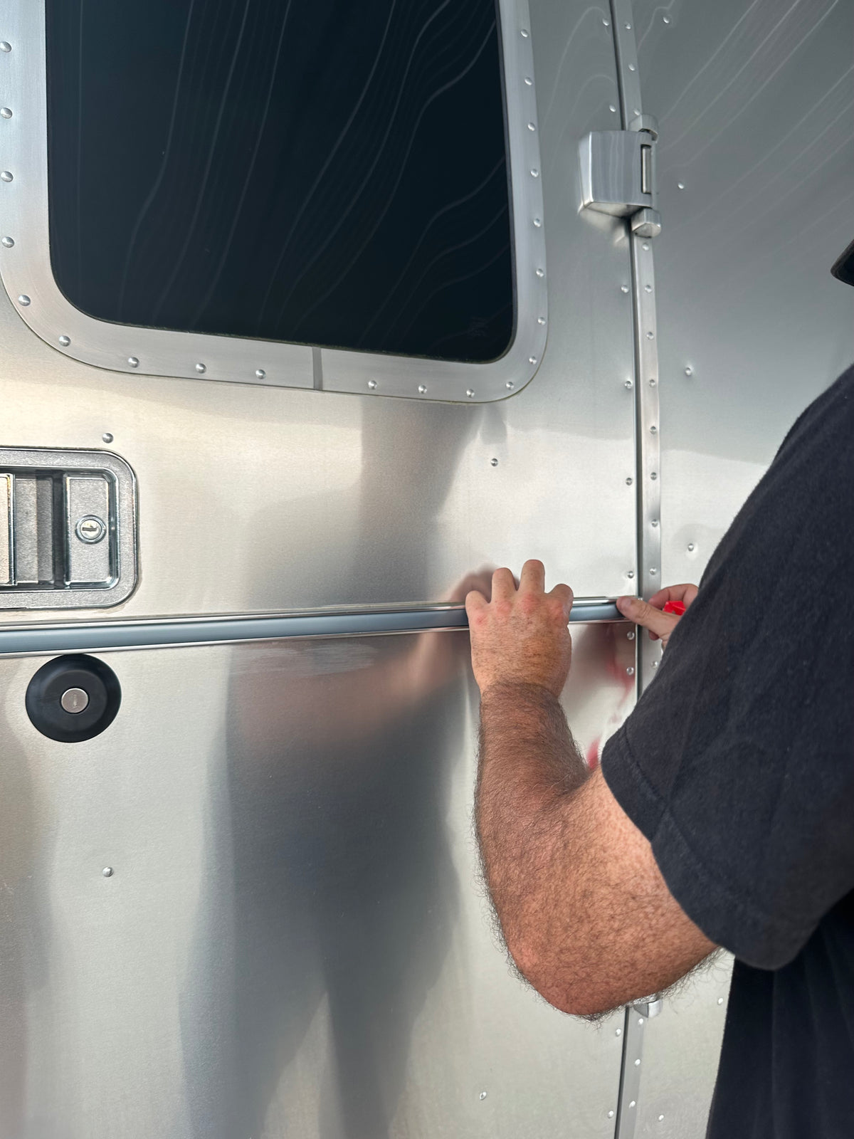 A person uses both hands to hold a metal bar against the shiny exterior of an Airstream trailer, likely installing the Airstream Premium Replacement Belt Trim for Travel Trailers—an essential exterior accessory—with part of their arm and a window visible.
