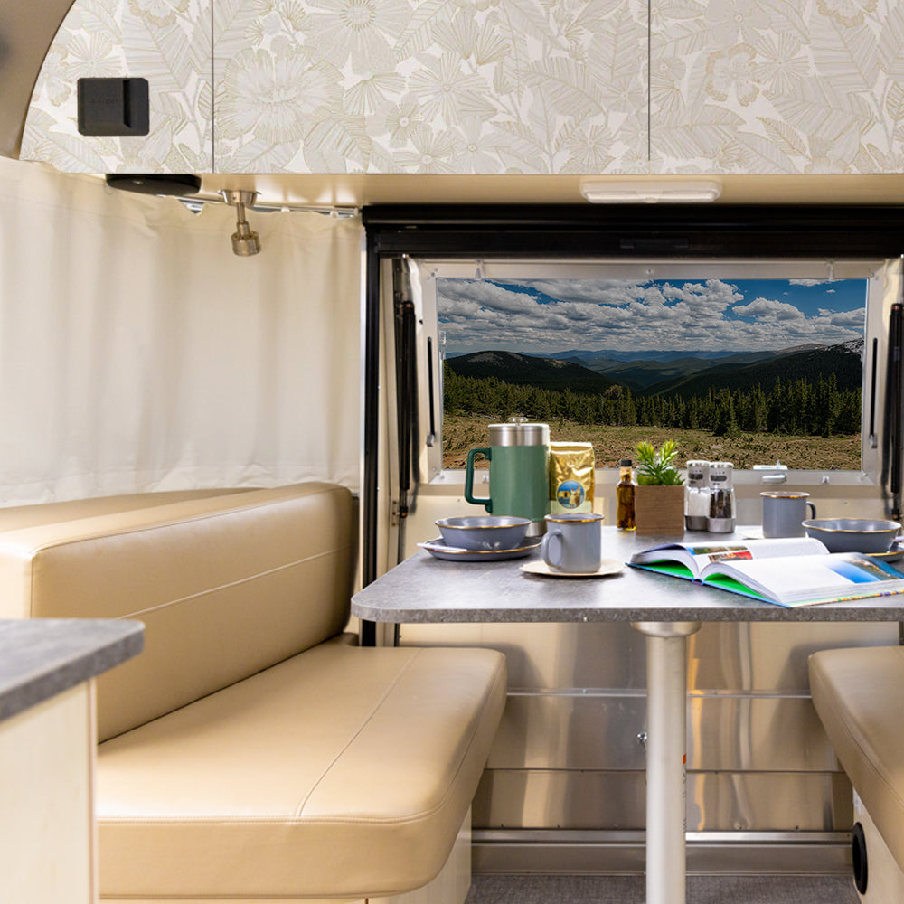A cozy RV dining area with tan seats and a set table, featuring the stylish Metallic Bloom Peel + Stick Removable Wallpaper that complements the inviting space, looking out through an open door to a scenic view of mountains, forest, and a cloudy sky.