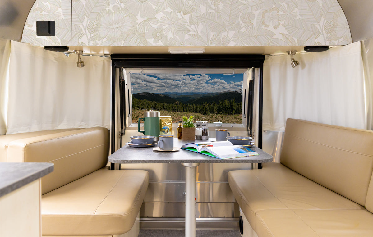 The interior of an Airstream features tan seating and a set table, where a book and coffee pot are thoughtfully arranged. A large rear window offers stunning views of rolling green hills, forest, and blue sky dotted with clouds. The walls are enhanced by the stylish Metallic Bloom Peel + Stick Removable Wallpaper, adding a modern touch to the inviting space.