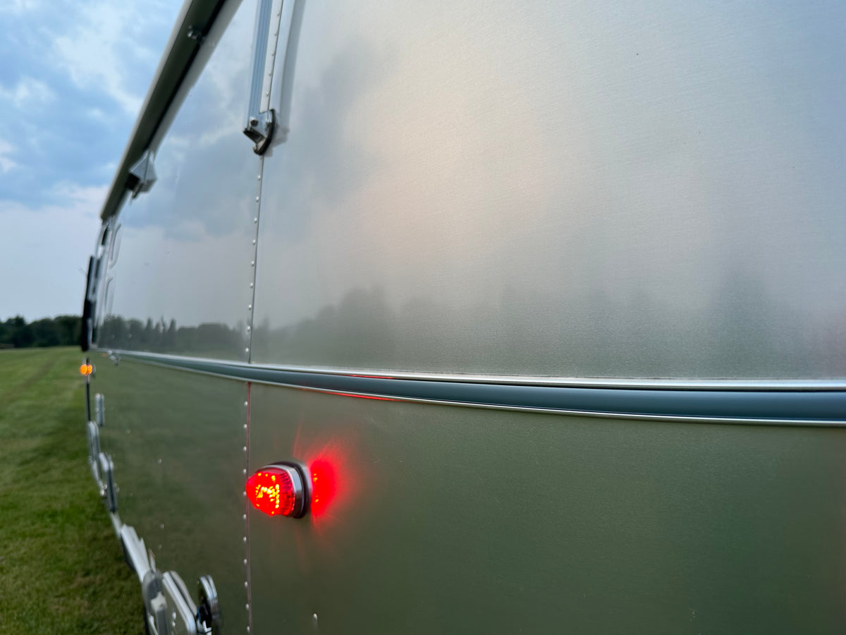 Close-up of a silver trailer with a red light illuminated, parked on grass beneath a cloudy sky. The sleek exterior is accented by the Airstream Premium Replacement Belt Trim for Travel Trailers, while trees are visible in the distance.