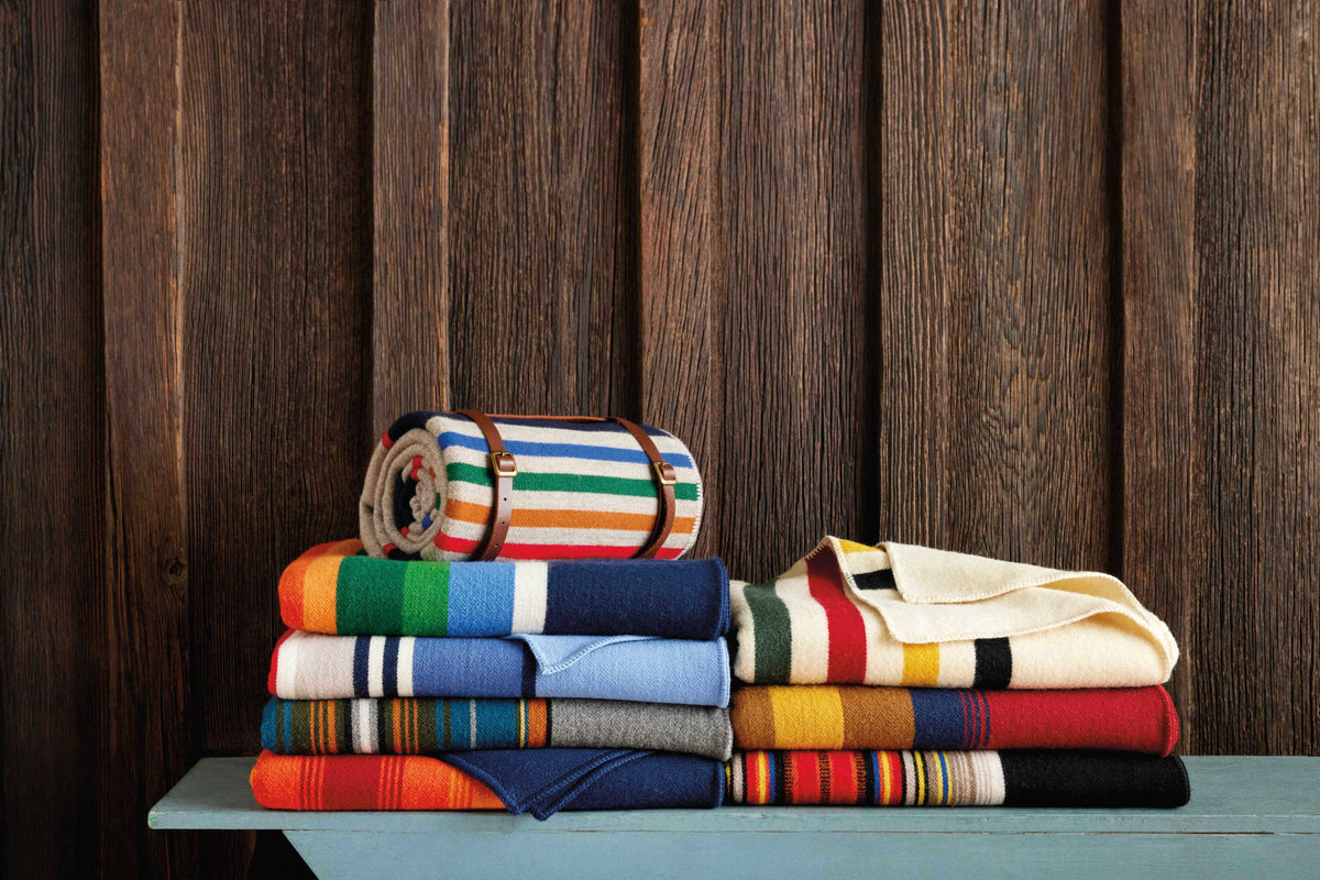 A stack of folded, colorful striped blankets, including a classic Pendleton wool throw, sits on a blue wooden bench with a rolled-up National Parks Throw with Carrier by Pendleton placed on top, all set against a rustic wooden wall background.