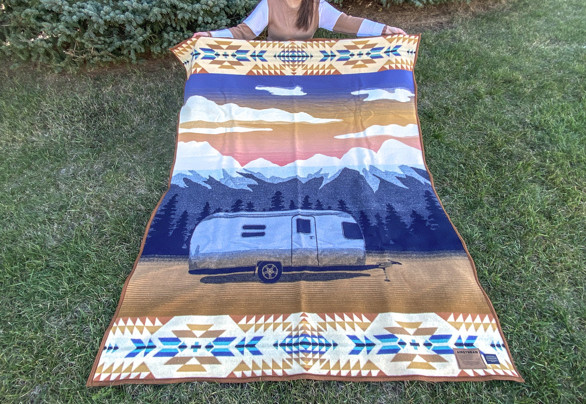 A person holds the Airstream x Pendleton Woolen Mills Jacquard Blanket outdoors. Inspired by Pendleton, the patterned blanket features a trailer set against mountain scenery with geometric designs bordering the top and bottom edges. Green grass and a tree appear in the background.
