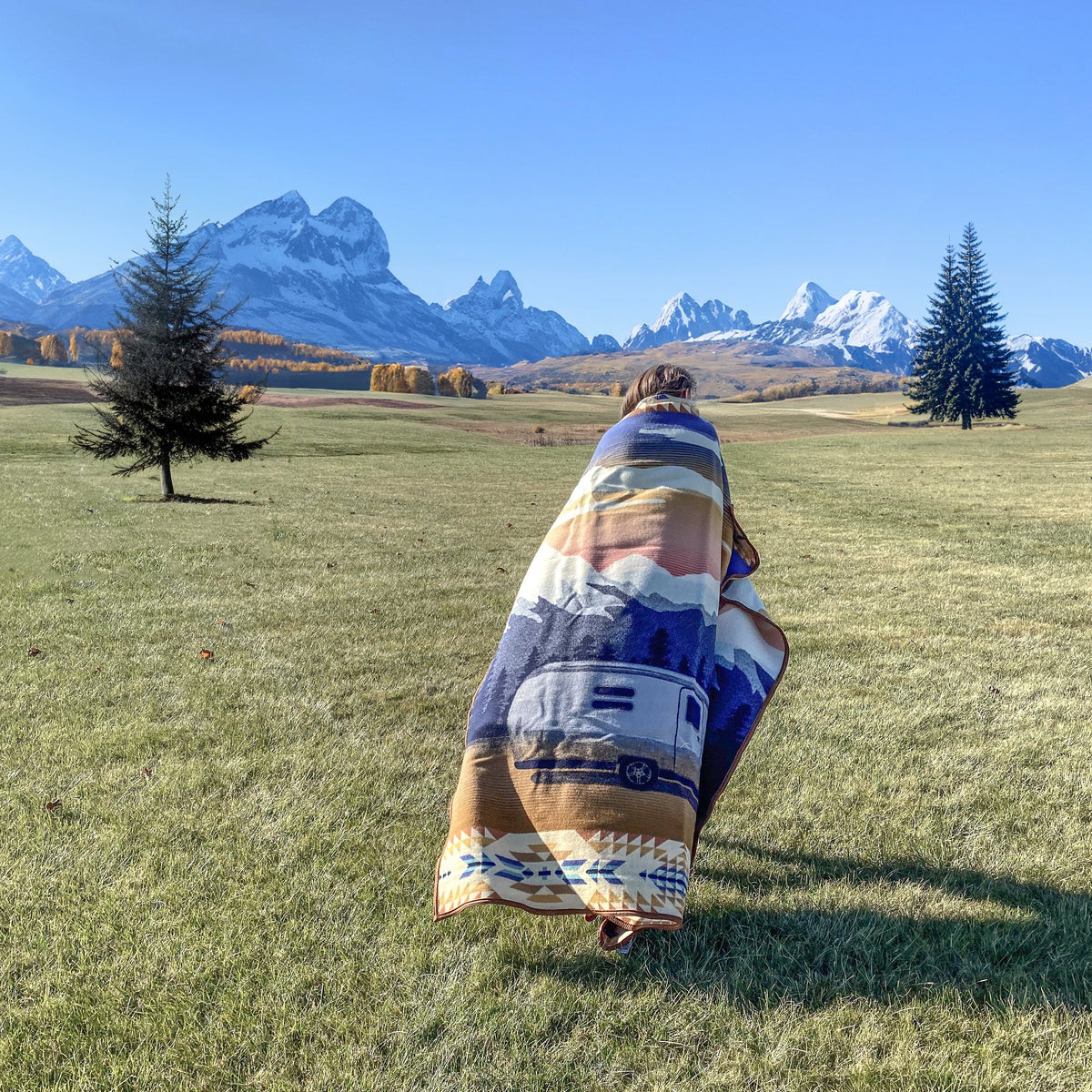 A person draped in an Airstream x Pendleton Woolen Mills Jacquard Blanket featuring mountain-inspired designs walks across a grassy field toward distant snow-capped mountains and evergreen trees beneath a clear blue sky.