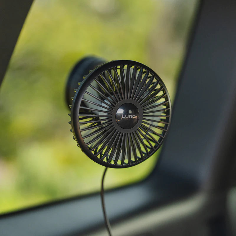 A small black USB Powered Camping Fan by Luno, featuring a circular grill and “Luno” written in the center, is attached to a car window—making it perfect for van life and car camping adventures. This USB-C powered fan is connected by a cord, with greenery visible just outside.