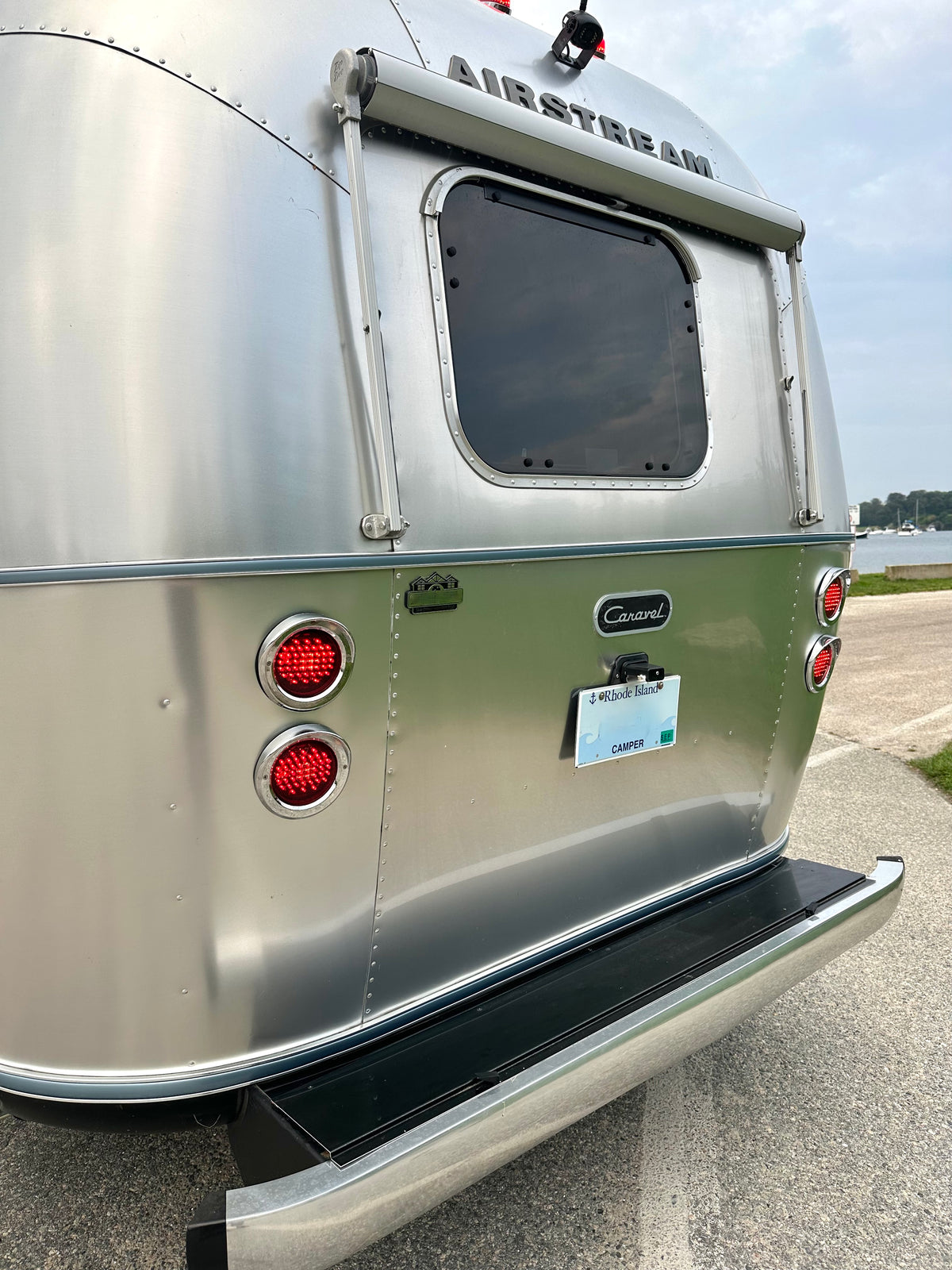 A silver, riveted aluminum Airstream travel trailer is shown from the rear, featuring red tail lights, a window, and a license plate reading “CAMPER.” This trailer is equipped with the Airstream Premium Replacement Rub Rail for Travel Trailers and is parked on a paved surface beside grassy terrain with water visible in the background.