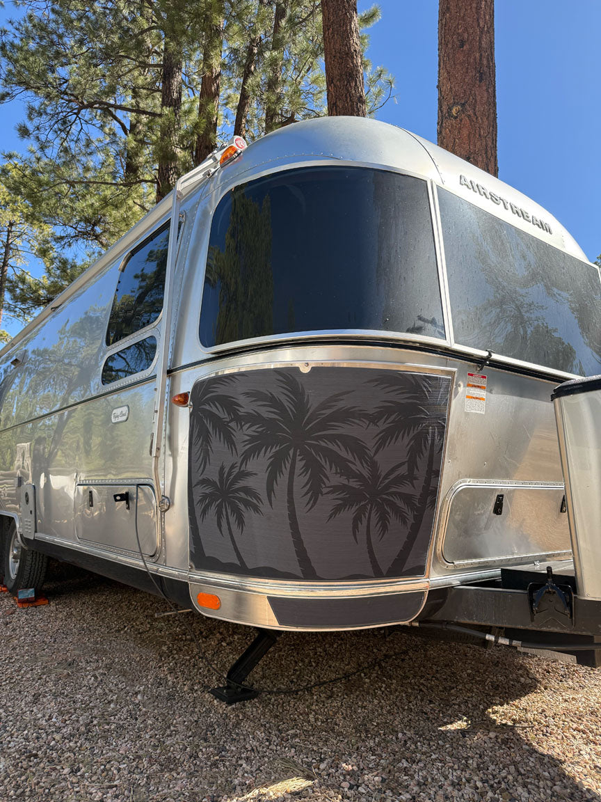 A silver Airstream trailer with palm tree graphics on the front is parked on gravel beneath tall pine trees, set against a clear blue sky. Its front features the Airstream Rock Guard Rock Wraps Nature Collection, offering durable protection for the travel trailer while enhancing its sleek appearance.