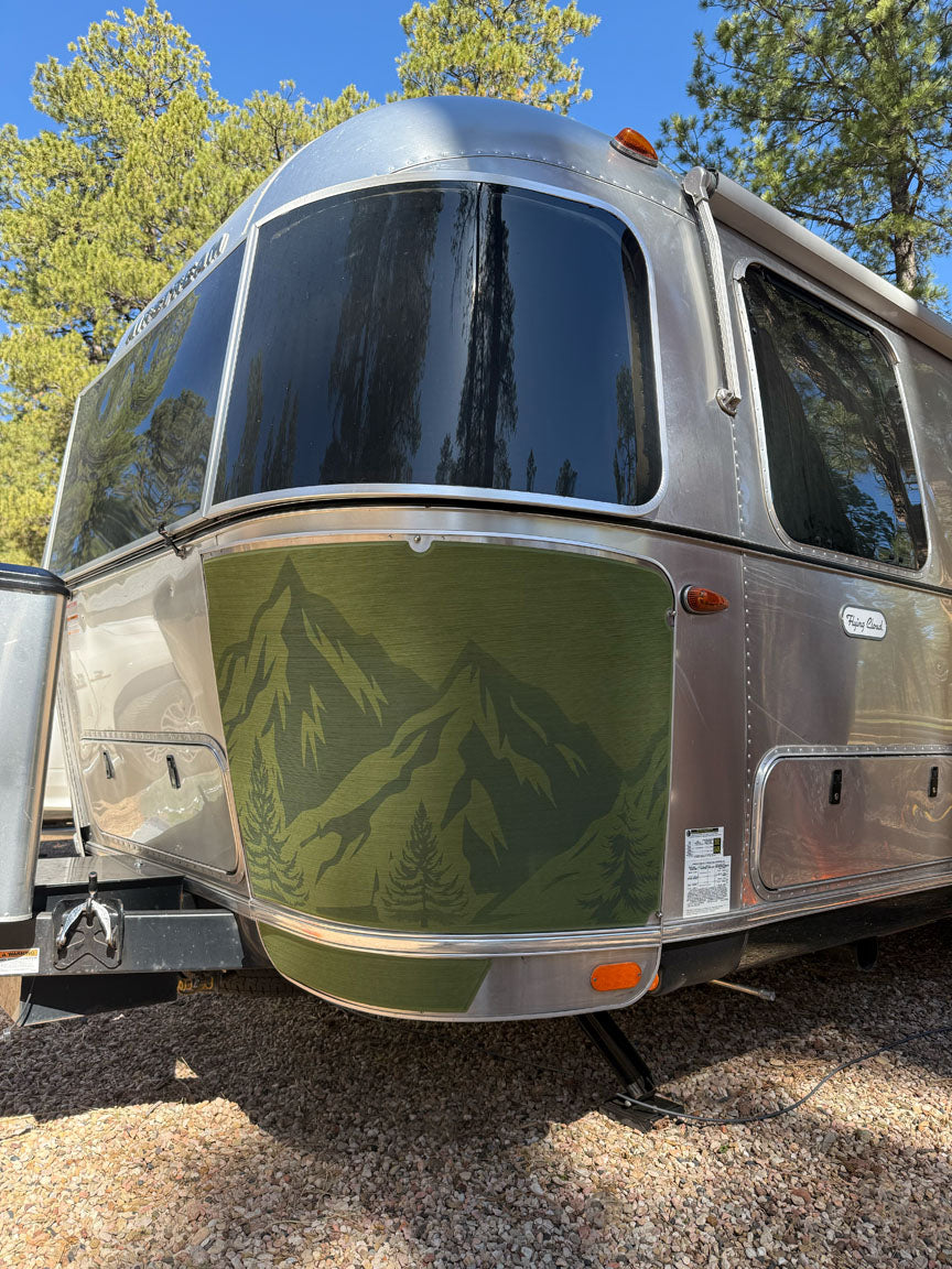 A silver travel trailer with a green mountain and tree mural on the front sits parked on gravel amidst tall pine trees under a clear blue sky. Its exterior features the Airstream Rock Guard Rock Wraps Nature Collection, providing enhanced protection and stylish durability for every adventure.