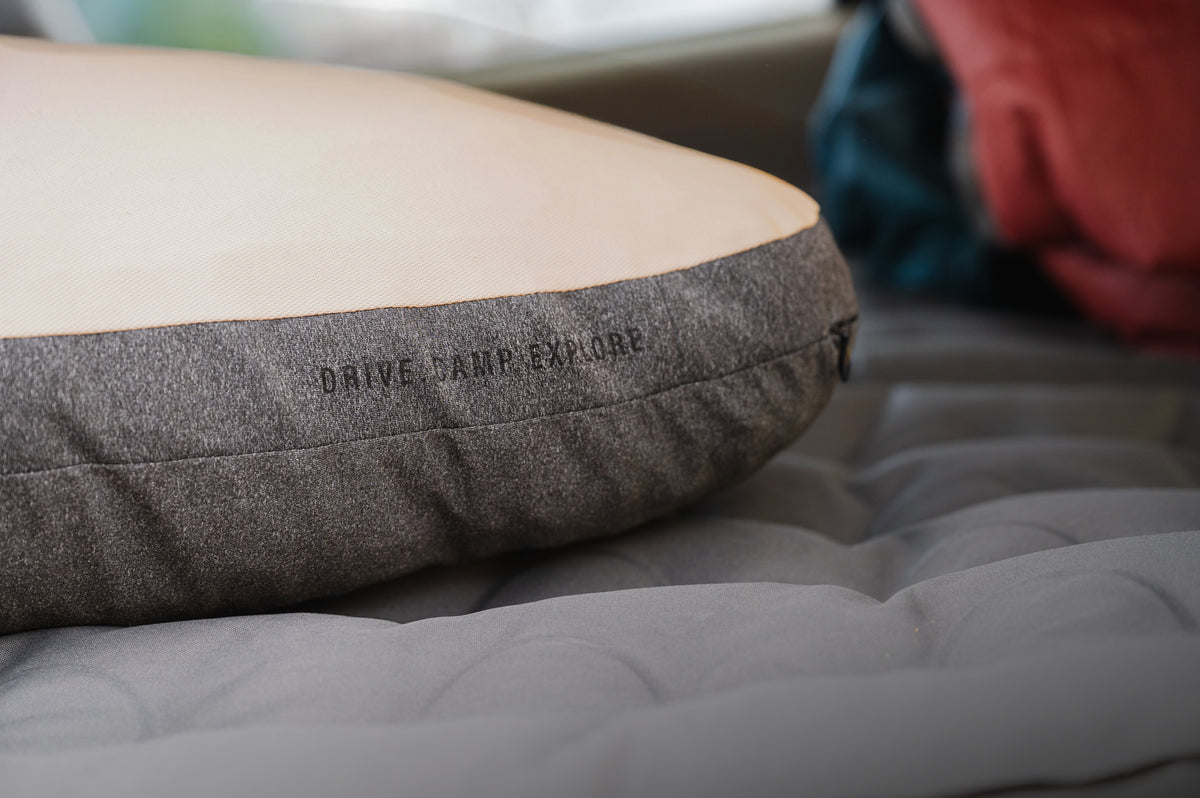 A close-up of the Packable Camp Pillow by Luno, featuring gray and beige tones with “DRIVE. CAMP. EXPLORE.” stitched on the side, resting on a gray sleeping pad—perfect for bringing Airstream gear style to your next adventure.