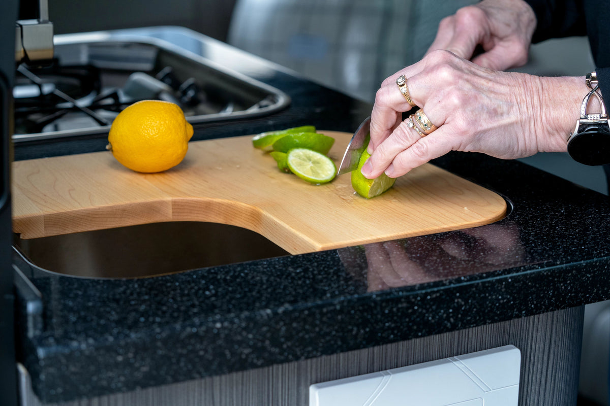 A person slices a lime on an Airstream Custom Sink Cutting Board for Interstate 24GT/GTX next to a whole lemon, in a modern kitchen with a black countertop.
