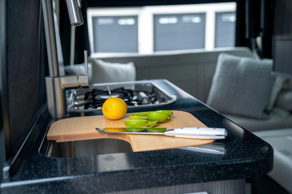 A cutting board with a knife, sliced lime, and a whole lemon sits on a black kitchen counter next to a sink and gas stove inside a modern RV or camper, featuring the Airstream Custom Sink Cutting Board for Interstate 24GT/GTX that adds both style and functionality to the space.