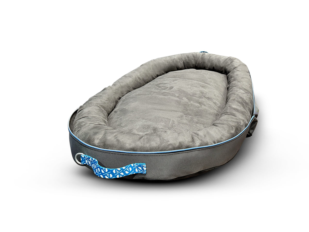Introducing the Airstream Travel Pet Bed: a grey, oval-shaped pet bed with a soft, plush cushion and raised sides for added comfort. Designed for both style and portability, it features a blue patterned handle on one side—perfect for travel or home use. The bed is shown against a white background.