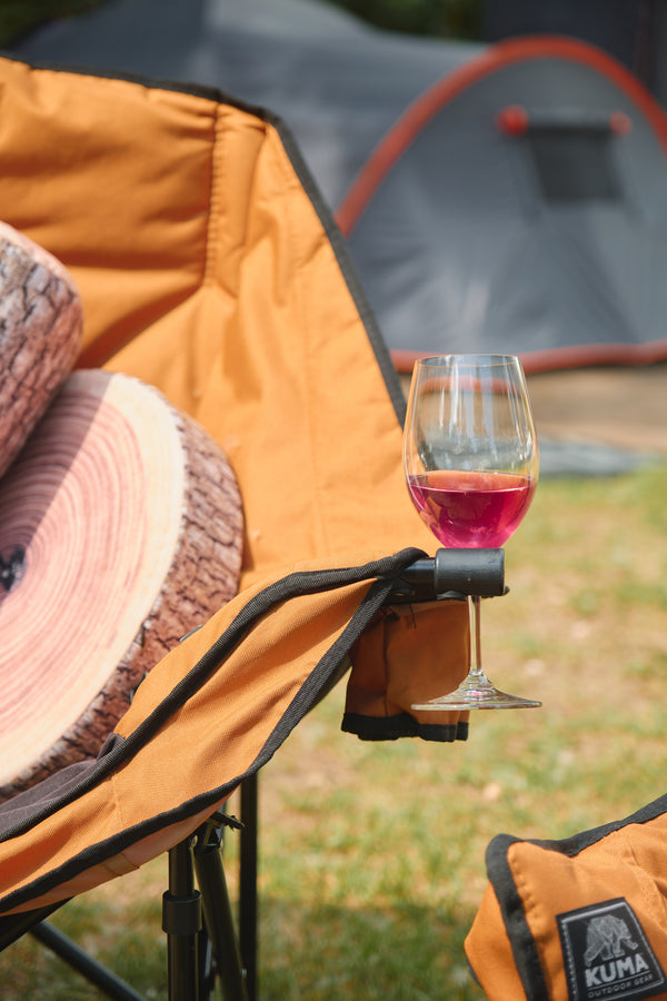 An orange camping chair with a built-in cup holder holding a glass of pink wine, set outdoors on grass with a tent and log slices in the background. A Kuma brand patch is visible on an orange bag in the lower right corner.