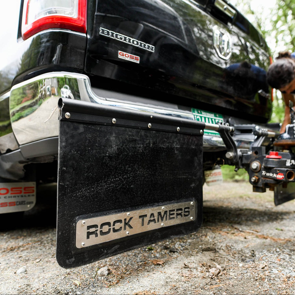 Close-up of a black truck’s rear with the Airstream Rock Tamers™ Mud Flap System providing debris protection and a trailer hitch attached, parked on a gravel surface. A person is partially visible adjusting equipment near the hitch.