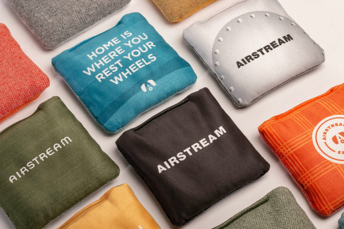 A set of Airstream ACA Cornhole Bags—colorful square fabric pouches featuring the AIRSTREAM logo and one with the phrase “HOME IS WHERE YOU REST YOUR WHEELS”—are arranged in a neat grid on a light surface.