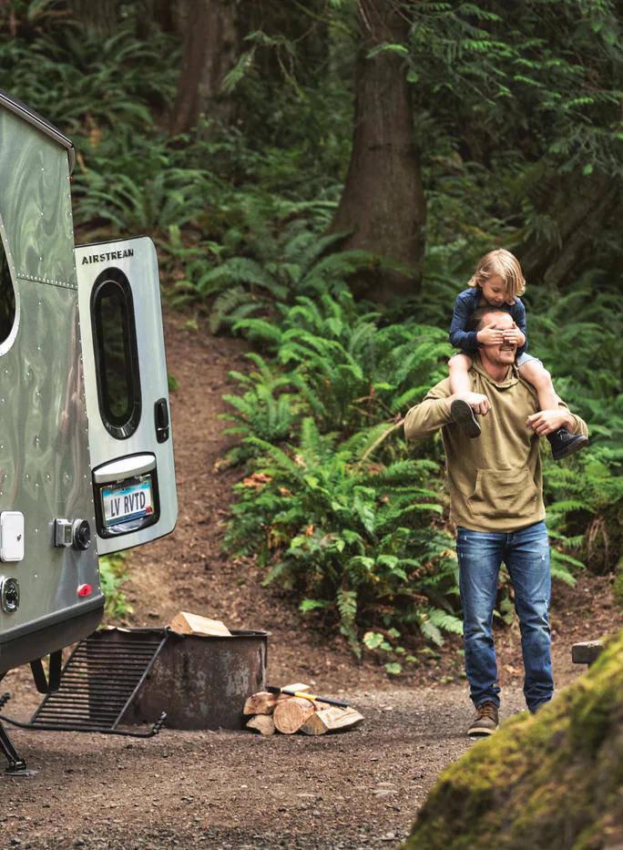 A man stands next to a camper in a forest, smiling with a child on his shoulders. Firewood and a fire pit are nearby, with green ferns and tall trees in the background.