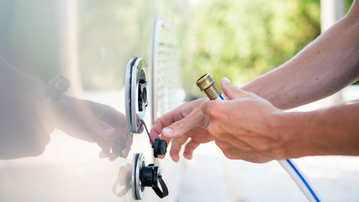 A person connects a hose to an outdoor water hookup on a white vehicle, possibly an RV, with a blurred green background.