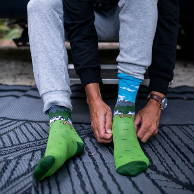 Wearing the Airstream Campfire Socks—one green with a sheep design and one blue featuring a mountain scene—a person dressed in gray sweatpants and a black sweatshirt sits on a mat, adjusting their cozy mismatched socks.