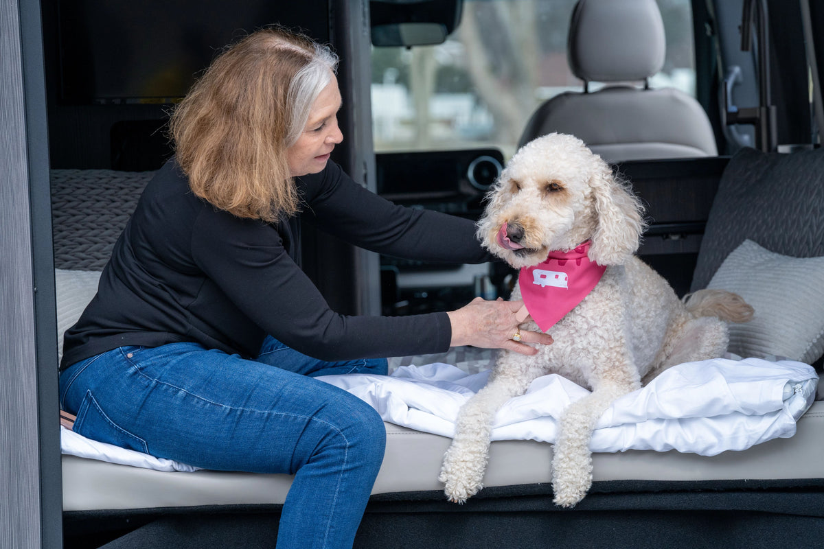 A woman sits on a bed in a van, petting a curly-haired dog wearing a pink bandana. Beneath her is the cozy Airstream x RVSuperbag All-In-One Bedding System, adding comfort to the scene as the dog sits calmly, looking forward and licking its lips.