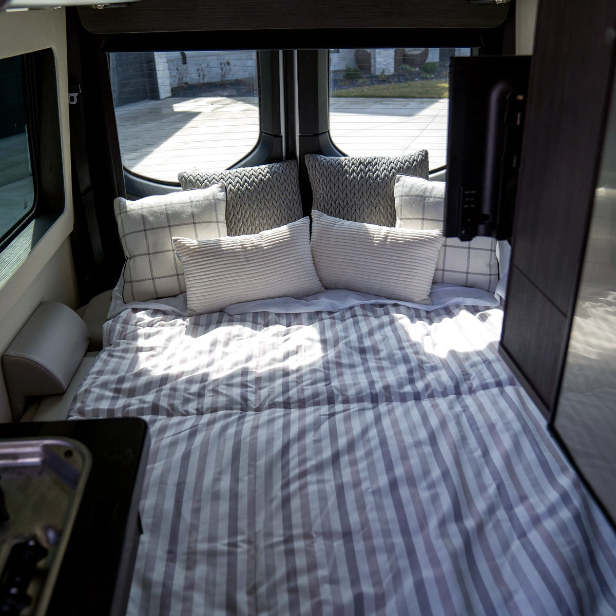 In an RV flooded with sunlight from expansive windows, a cozy bed is adorned with striped bedding and a mix of patterned pillows. The space features the Airstream x RVSuperbag All-In-One Bedding System, specifically crafted for comfort on the road, adding to the modern and inviting interior.