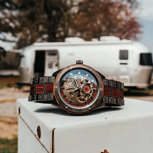 Luxury wristwatch with intricate design on a white surface, blurred background of trees and RV.