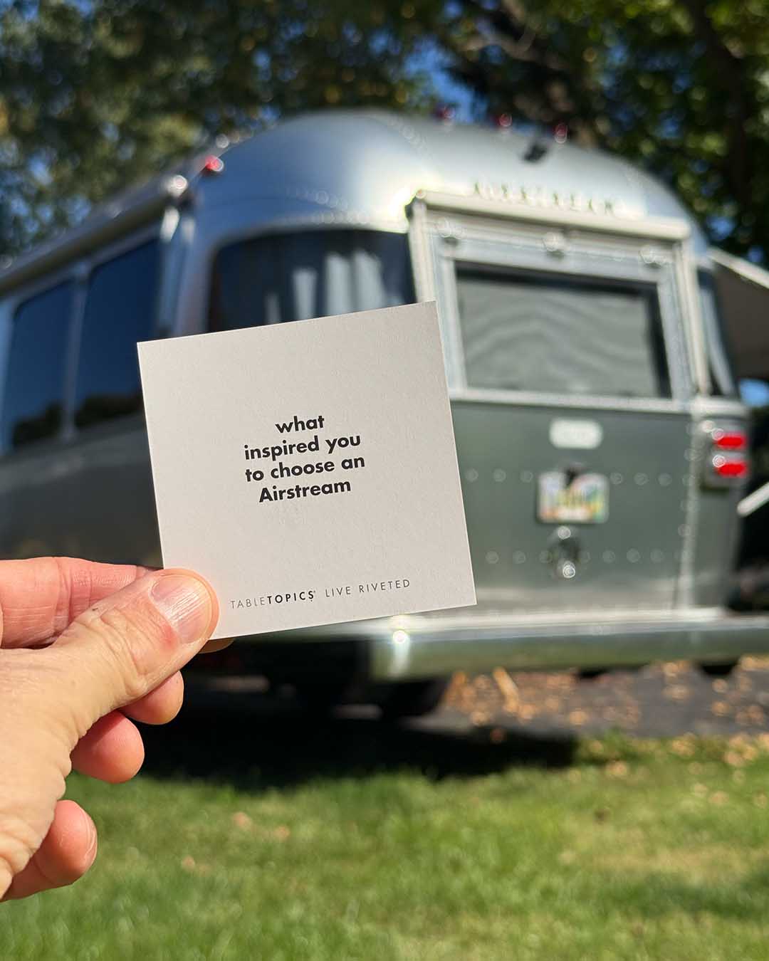 A hand holds a square Airstream TableTopics Game card asking about the inspiration behind choosing the trailer, held in front of a parked silver camper.