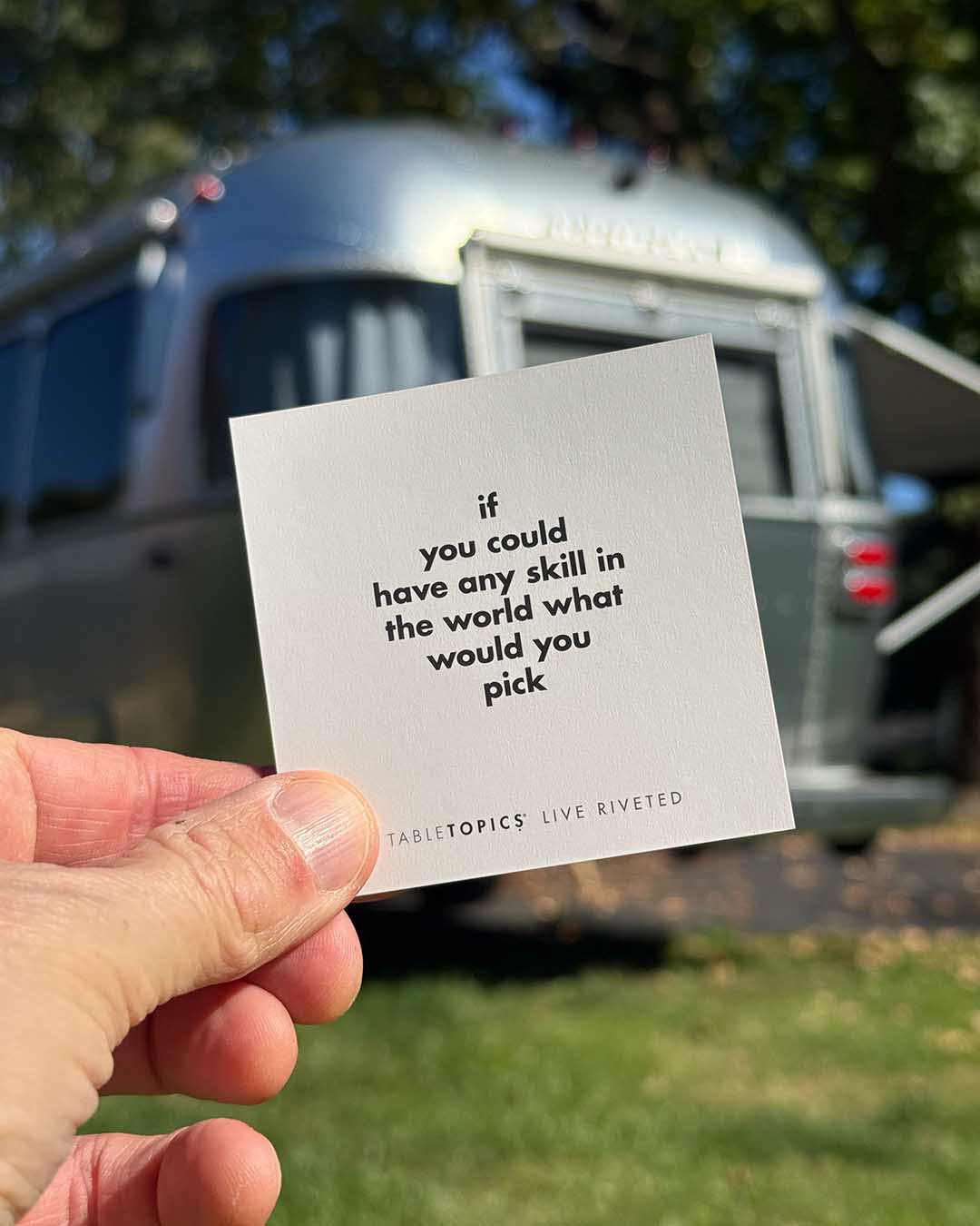 A hand holds an Airstream TableTopics Game card that reads, If you could have any skill in the world what would you pick, in front of a parked travel trailer.