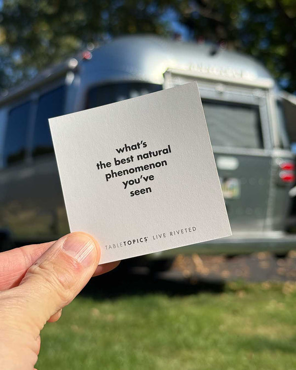 A hand holds a square Airstream TableTopics Game card that reads, what’s the best natural phenomenon you’ve seen, with a travel trailer blurred in the background.
