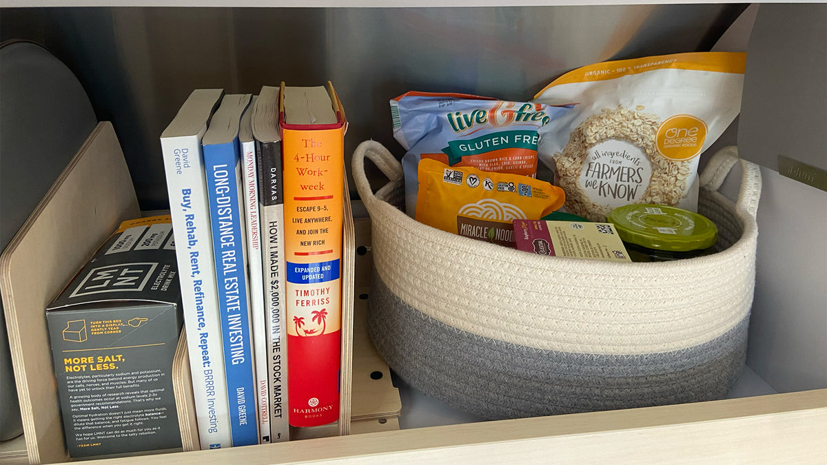 Books, including *The 4-Hour Work Week*, are arranged beside a woven basket filled with snacks like popped chips, rice cakes, and guacamole cups on a shelf enhanced by the Airstream Touring Coach Roof Locker Organization from Airworthy Equipment Company for optimal modular roof locker organization.