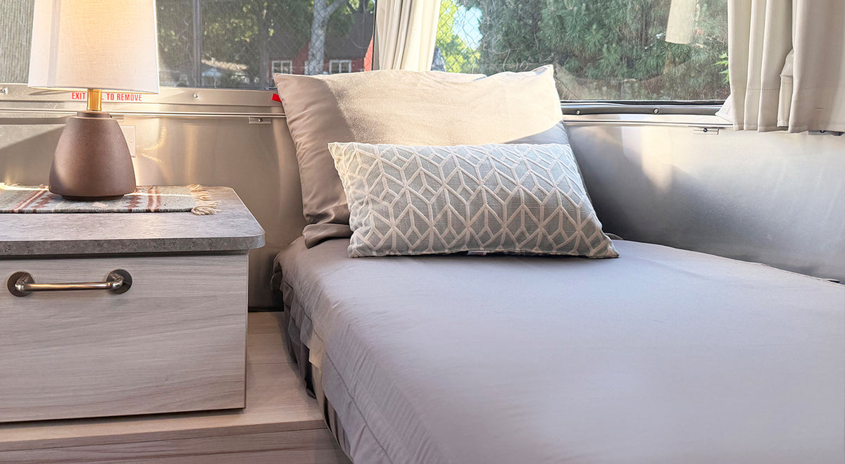 A neatly made bed with the Airstream Custom Fit Beddy’s ZipBase Sheet Set for Trade Wind Travel Trailers in gray and a patterned pillow sits next to a small nightstand with a lamp in a cozy, sunlit room.