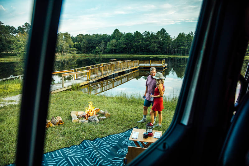A man and woman stand near a campfire by a lake with a wooden dock, surrounded by trees. Photo is taken from inside a vehicle, with a picnic blanket and supplies on the grass.