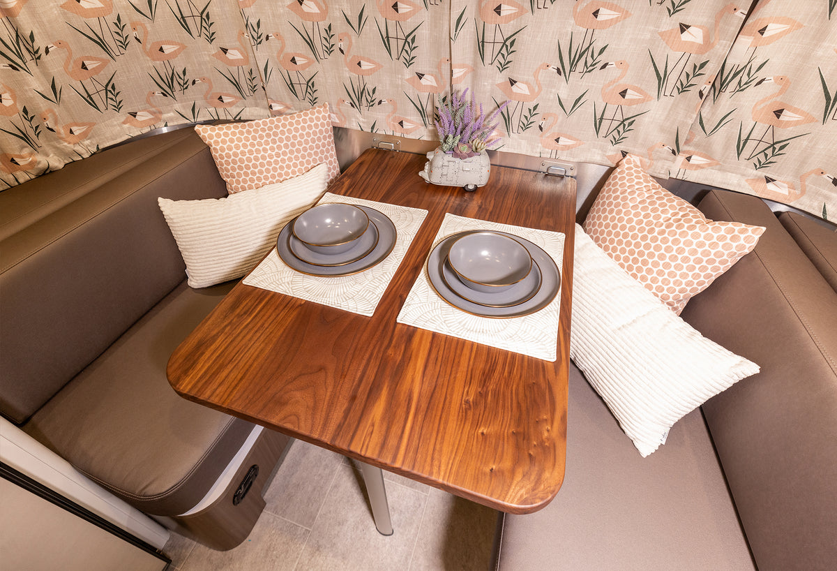 The Airstream Wood Dinette Table Upgrade for Flying Cloud Travel Trailers is shown in an RV with two place settings, brown cushions, patterned and white pillows, and beige floral curtains in the background. A small decorative plant sits at the center of this premium wood dinette table.