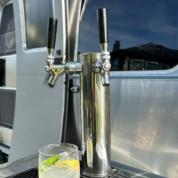 Metallic beer tap with two handles on a reflective surface, possibly a truck or van, with a glass of lemonade in the foreground.