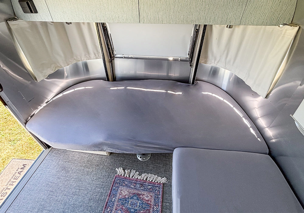 A modern RV interior features a neatly made bed dressed with Airstream Dinette Fitted Sheets for Caravel Travel Trailers, complemented by a small rug on the floor, white curtains, and cabinets above.