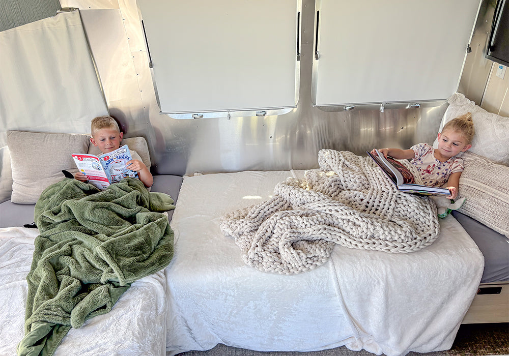 Two young children are lying on separate beds inside a camper, each reading a book. They are covered with cozy blankets—one green and one chunky beige—creating a comfortable, relaxed atmosphere. The beds are dressed with Airstream Dinette Lounge Cushion Fitted Sheets for Travel Trailers, custom-fit to provide extra comfort and style to their cozy setup.