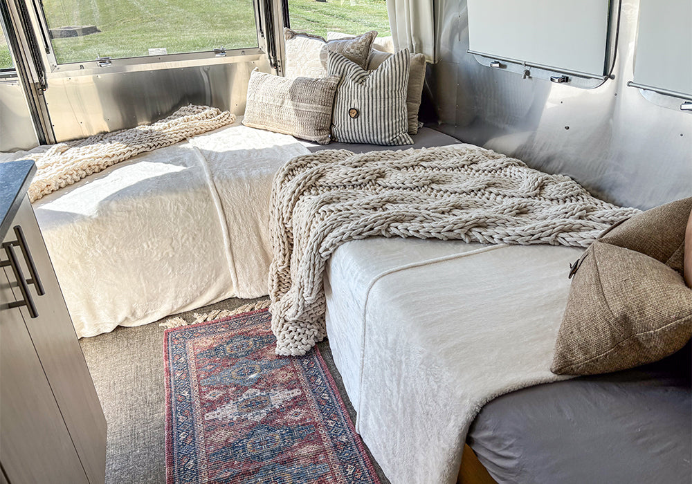 Cozy camper bedroom with two beds featuring Airstream Dinette Fitted Sheets for Victorinox Travel Trailers, a textured throw blanket, patterned rug, and decorative pillows next to large windows with a view of grass outside.