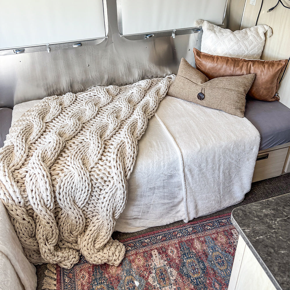 A cozy bed adorned with a chunky knit cream blanket and textured leather-and-fabric pillows sits against a metallic wall atop a patterned rug—perfectly complemented by the Airstream Dinette Lounge Cushion Fitted Sheet for Travel Trailers, designed to enhance both comfort and style in your unique space.