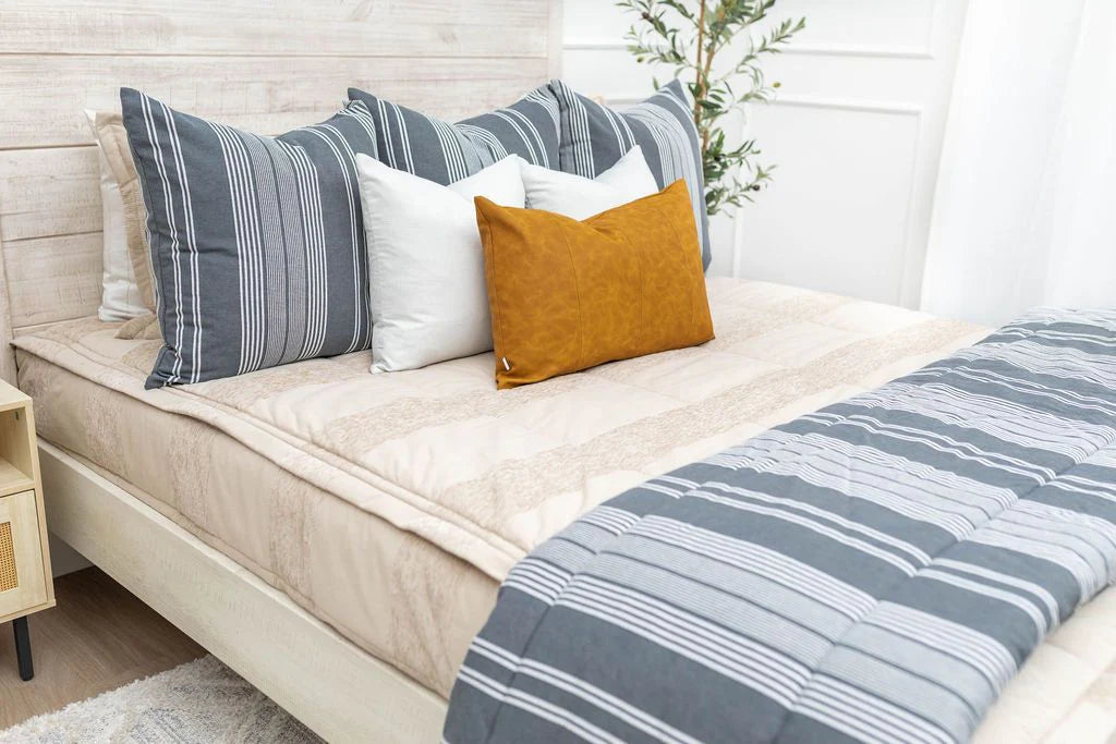 A neatly made bed outfitted with the Airstream RV Fit Beddy’s for Excella Travel Trailers in a bright, modern bedroom, featuring striped gray and white zippered bedding, a variety of pillows—including white, gray, and mustard yellow—and a small plant in the corner.