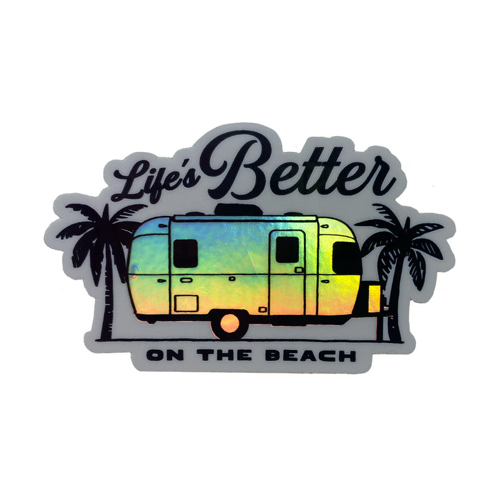 The Airstream Beach Holographic Trailer Sticker showcases a holographic camper trailer nestled between two palm trees, with the phrase “Life’s Better on the Beach” in stylized black lettering above and below the trailer.