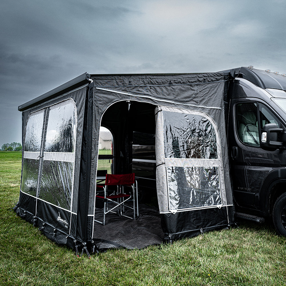 A black Airstream Rangeline campervan is set up on grass under a cloudy sky with the Buddy Awning Enclosure attached, featuring panoramic windows made from premium Isacryl material. Inside the enclosure are two folding chairs and a small table.