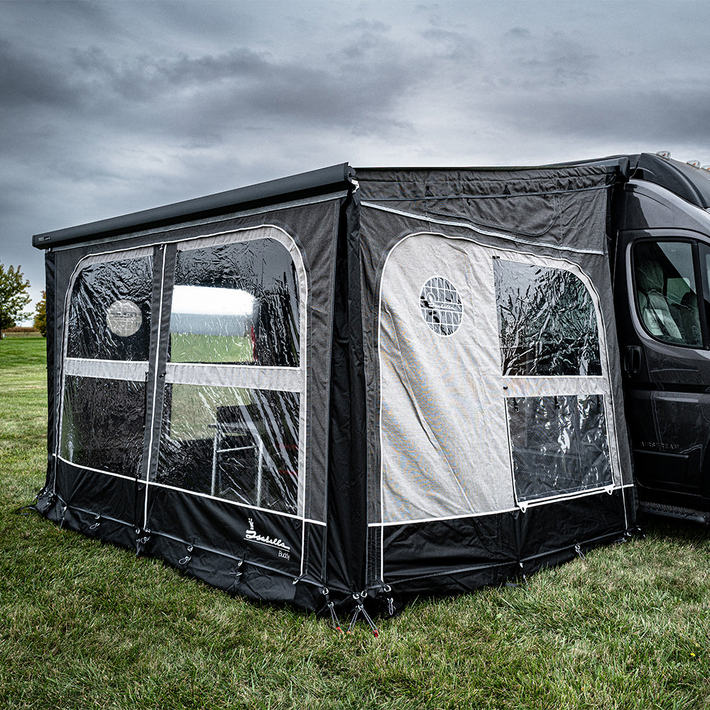 A black and gray Buddy Awning Enclosure for Airstream Rangeline, crafted from premium Isacryl material, is attached to a van and set up on a grassy field under a cloudy sky. The enclosure features large panoramic windows and a door flap, providing an enclosed outdoor space.