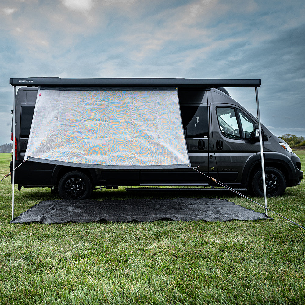 A black camper van is parked on grass with its awning extended. The Shade Screen for Airstream Rangeline, a white side panel serving as a canopy shade screen, is attached to the awning to provide sun protection and privacy. A cloudy sky is visible in the background.